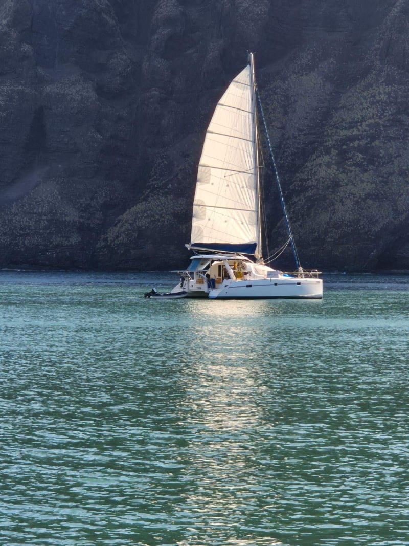 Le catamaran Toake Ona sous grand-voile devant une falaise